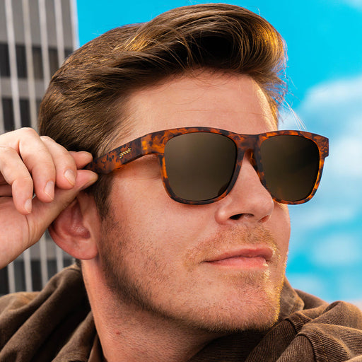 A person wearing wide-fit brown tortoiseshell sunglasses with non-reflective brown lenses looks to the side, confident.